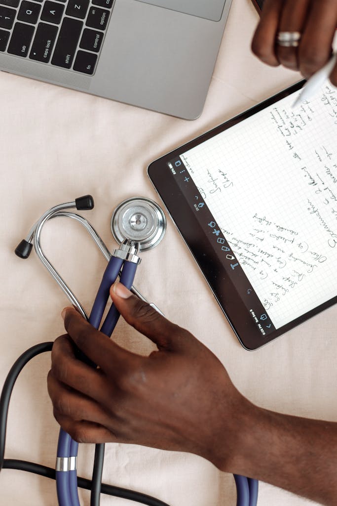 A close look at hands holding a stethoscope next to a tablet showing medical notes.